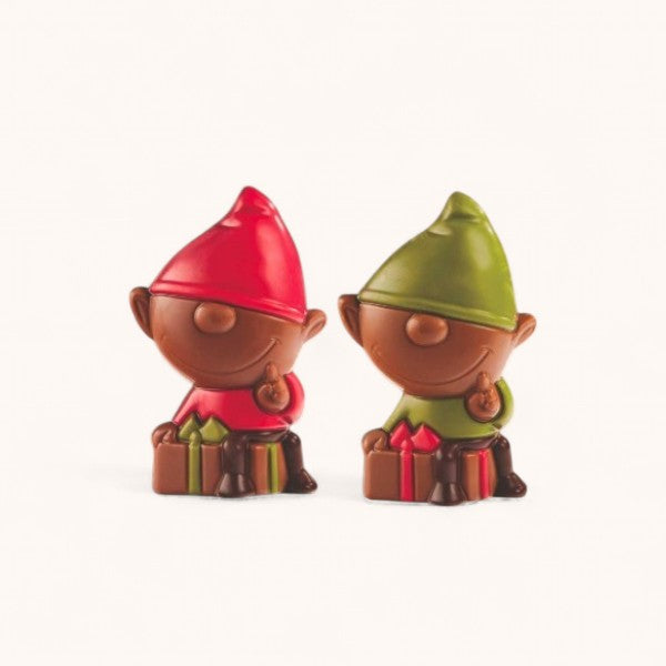 Deux figurines en chocolat en forme de lutins de Noël, l’un avec un bonnet rouge et l’autre avec un bonnet vert, assis à côté de petits cadeaux colorés.