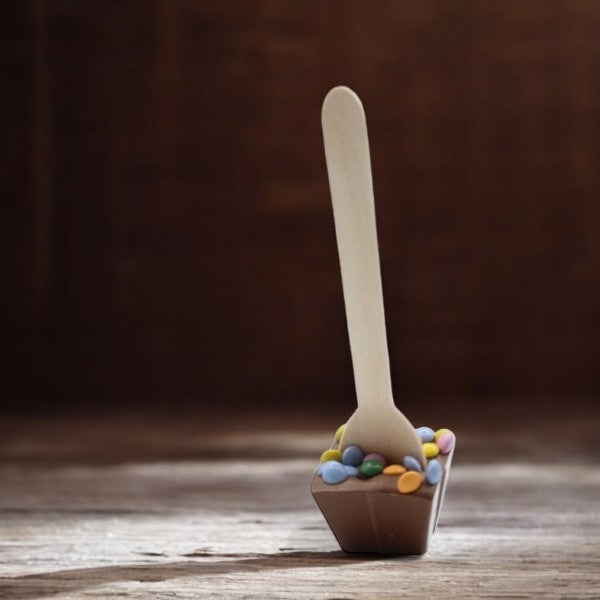 Cuillère à chocolat chaud au lait et pastilles colorées sur fond bois – gourmandise fondante et festive à déguster.