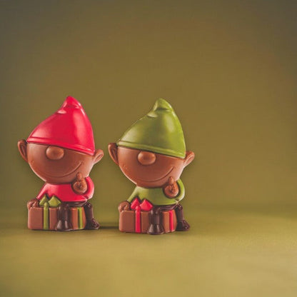 Deux lutins de Noël en chocolat au lait posés sur fond vert, moulages gourmands et colorés pour les fêtes.