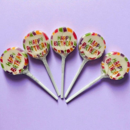 Cinq sucettes Rock “Happy Birthday” sur fond violet – confiseries colorées au cassis pour fête d’anniversaire.