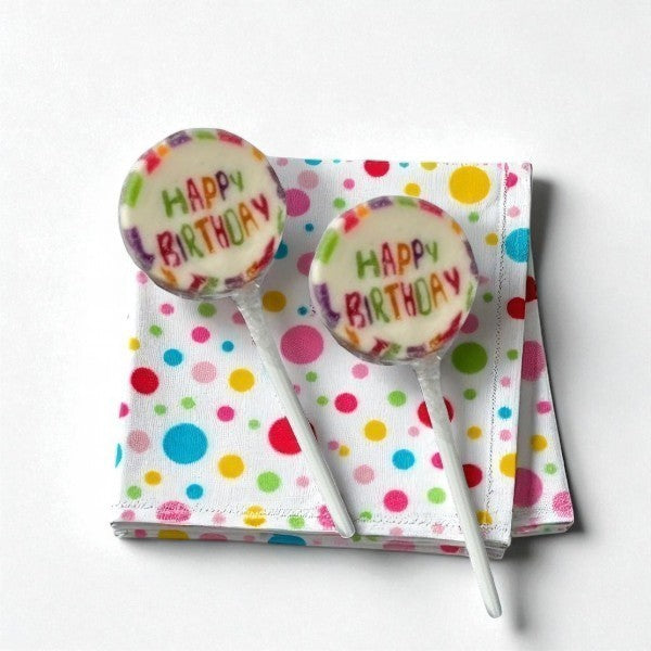 Deux sucettes Rock “Happy Birthday” posées sur une serviette d’anniversaire – bonbons festifs au cassis.