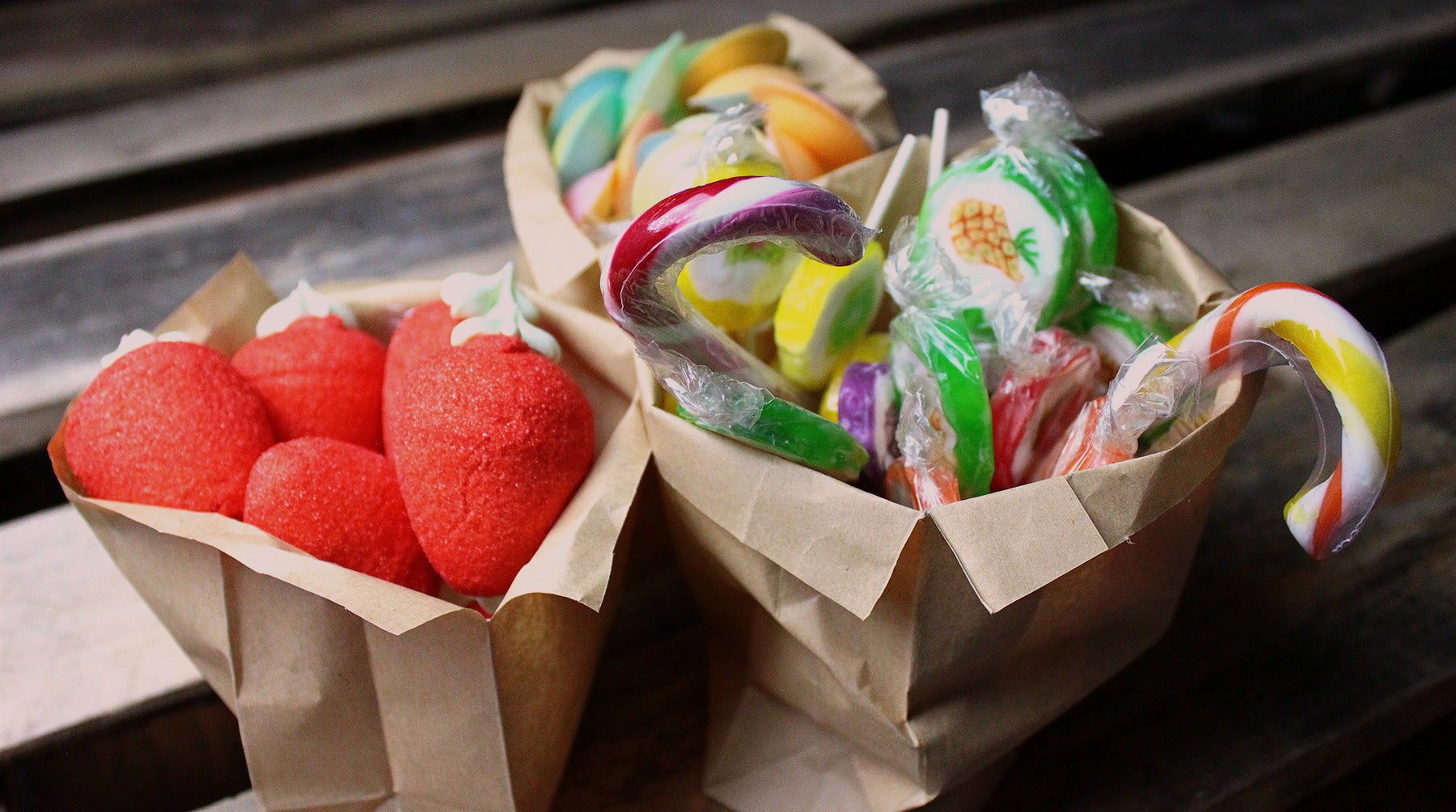 Trois sachets kraft de bonbons artisanaux posés sur un fond en bois, contenant des fraises géantes en guimauve rouge, des cannes à sucre colorées et une sucette rock multicolore.